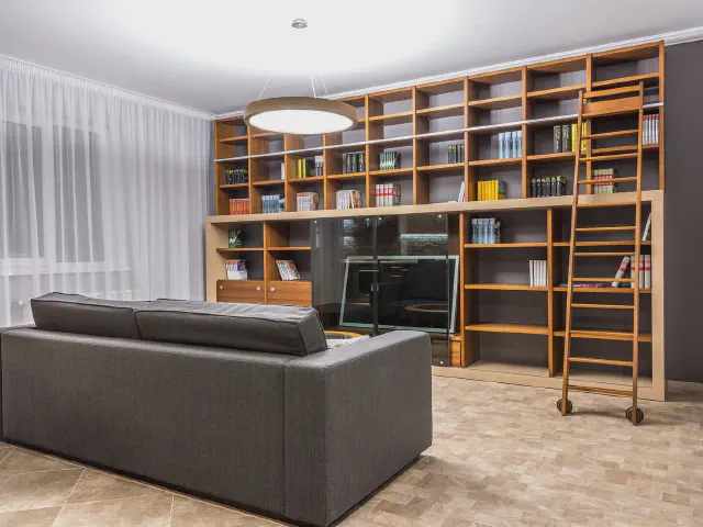 Salon z grafitową sofą i regałem na całą ścianę w kolorze drewna, z książkami, wnęką na telewizor i drewnianą drabiną na szynie