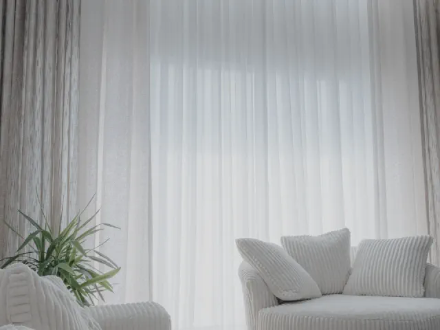 Białe, zwiewne firany z woalu do samej podłogi przy dużym oknie, obok szara zasłona, jasna sofa z poduszkami w prążki i roślina