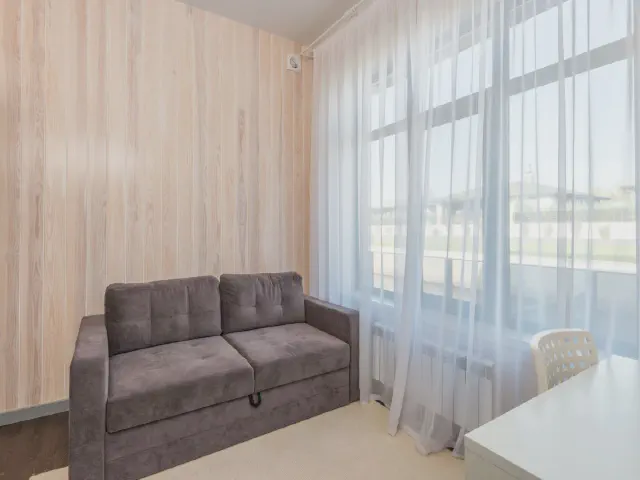 Szeroki salon z oknem na całą ścianę zasłoniętym przejrzystą białą firaną, szara sofa przy ścianie z jasnych desek i białe biurko