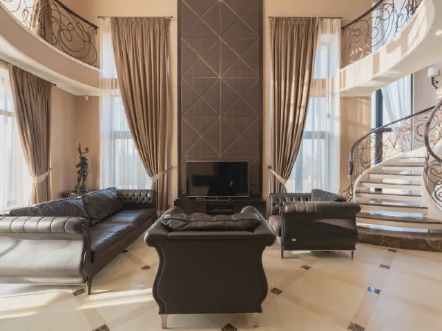Wysoki salon z antresolą i kręconymi schodami, przy wysokich oknach długie zasłony w kolorze taupe spływające do podłogi, czarne skórzane pikowane sofy