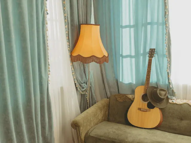 Miętowe zasłony z ozdobną lamówką i białe firany przy oknie, obok oliwkowa sofa, lampa z abażurem z frędzlami i gitara