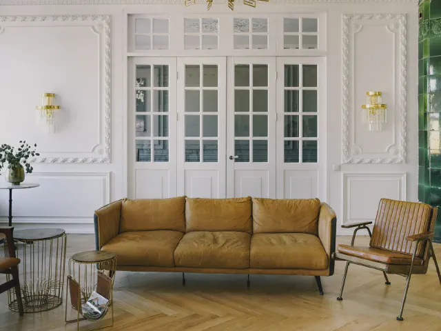 Salon z białymi drzwiami ze szprosami i sztukaterią na ścianach, koniakowa skórzana sofa, zielony kaflowy piec i jasny parkiet w jodełkę