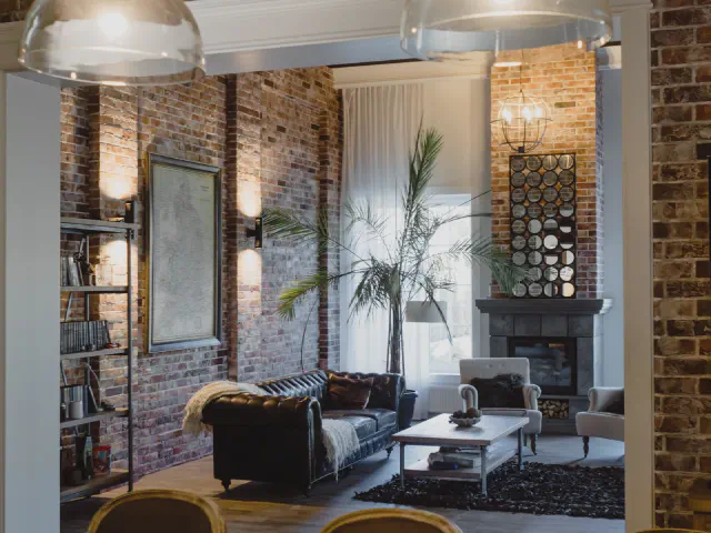 Wysoki salon w stylu loft widziany z jadalni: ceglane ściany z kinkietami, czarna skórzana sofa chesterfield, kominek i metalowy regał