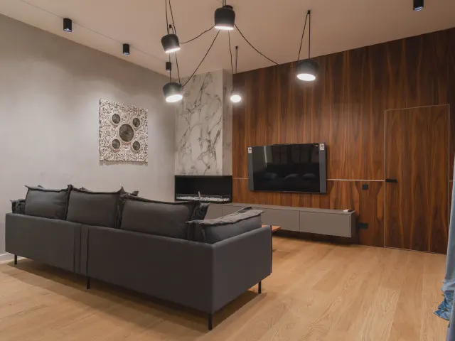Nowoczesny salon z grafitowym narożnikiem, ścianą telewizyjną wykończoną ciemnym drewnem o pionowym rysunku słojów, kominkiem w jasnej okładzinie i dębową podłogą