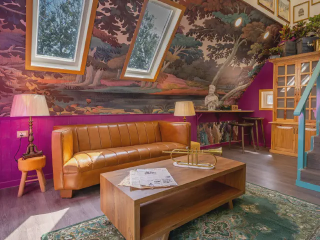 Salon na poddaszu z oknami dachowymi, ściana i skos pokryte motywem leśnego krajobrazu, koniakowa skórzana sofa, drewniana ława i ściana w kolorze fuksji