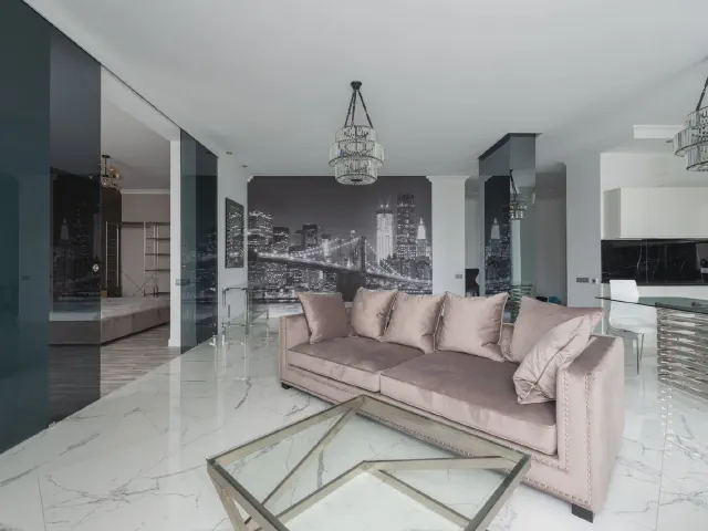 Jasny salon z marmurową podłogą, pudroworóżową sofą i szklanym stolikiem, na ścianie w głębi czarno-biała fototapeta z nocną panoramą miasta i mostem
