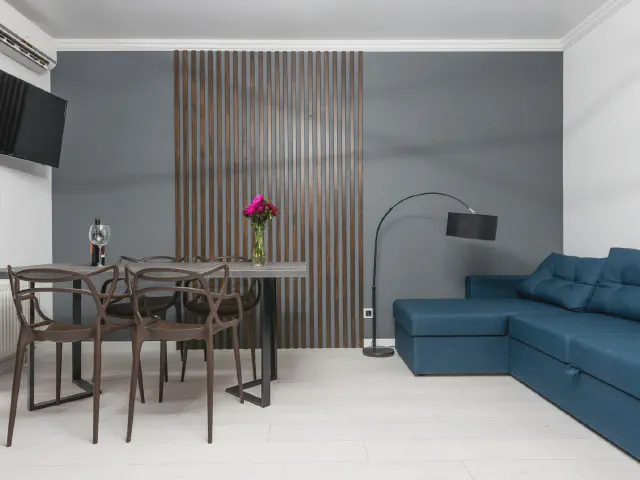 Salon z jadalnią, pas ciemnych lameli na szarej ścianie za stołem z krzesłami, obok granatowy narożnik i lampa łukowa