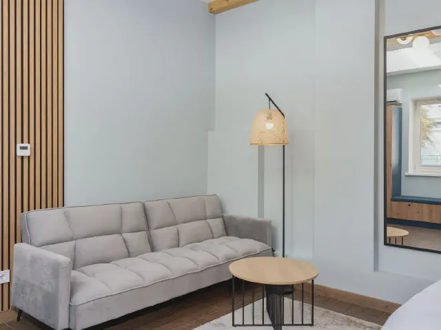 Jasnoszara sofa, okrągły stolik i lampa podłogowa z jasnym kloszem obok pasa drewnianych lameli na jasnej ścianie