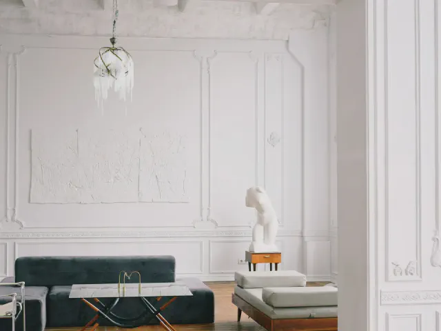 Biały salon ze ścianami podzielonymi prostokątnymi ramami ze sztukaterii, ciemnozielona sofa, szklany stolik, rzeźba na postumencie i kryształowy żyrandol