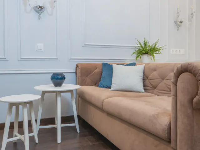 Karmelowa pikowana sofa z niebieską i białą poduszką przed jasną ścianą z ramami z listew, obok dwa białe stoliki i roślina na ścianie