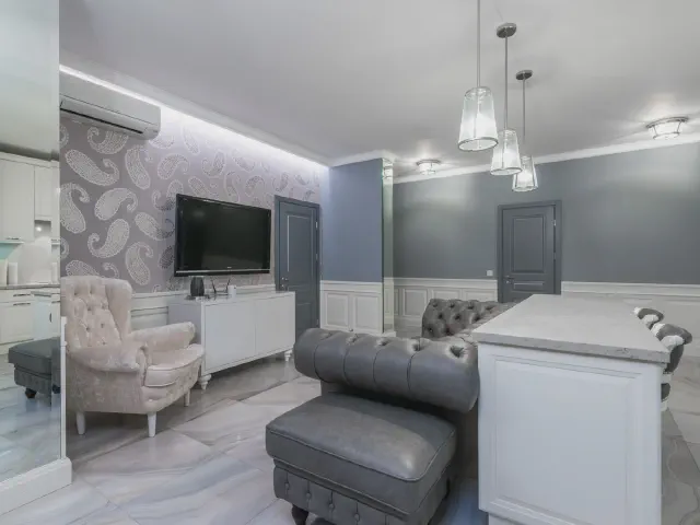 Salon z aneksem, ściana z tapetą w szary wzór paisley za białą szafką RTV z telewizorem, grafitowa ściana obok, pikowany fotel i skórzana sofa