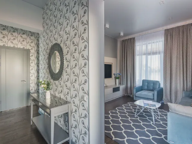 Wejście do salonu z pasem tapety w pnące, roślinne wzory na tle bieli, okrągłe lustro nad konsolą, w głębi niebieski fotel i dywan w geometryczny wzór
