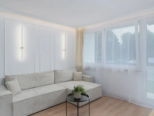Jasny minimalistyczny salon w bloku z białą sofą narożną, pionowymi kinkietami LED i dużym oknem