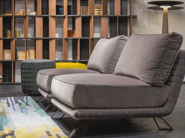 Nowoczesna szara sofa na płozach obok regału z drewnianymi półkami i pufami
