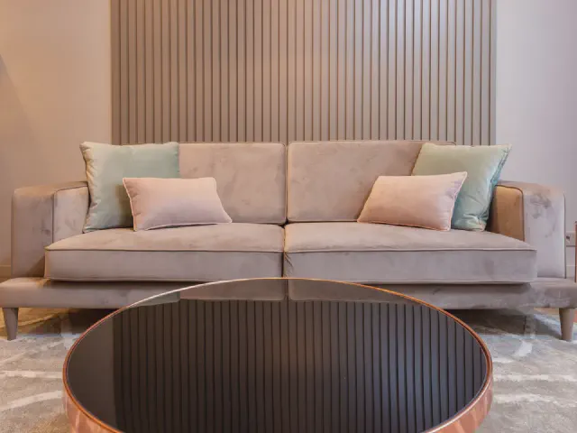 Beżowa sofa z pastelowymi poduszkami na tle lameli i okrągły stolik z miedzianą obręczą