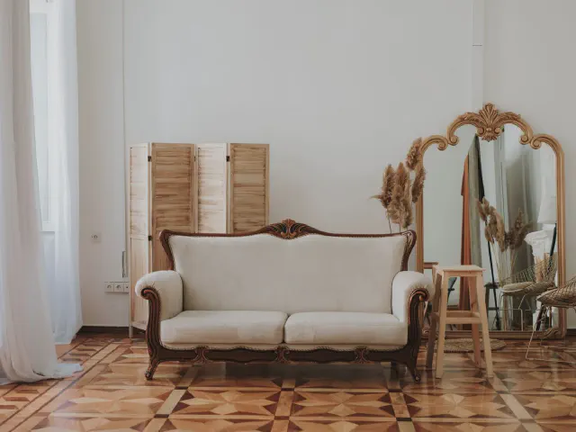 Stylizowana sofa w stylu Ludwika na parkiecie ułożonym we wzór, obok złocone lustro i drewniany parawan