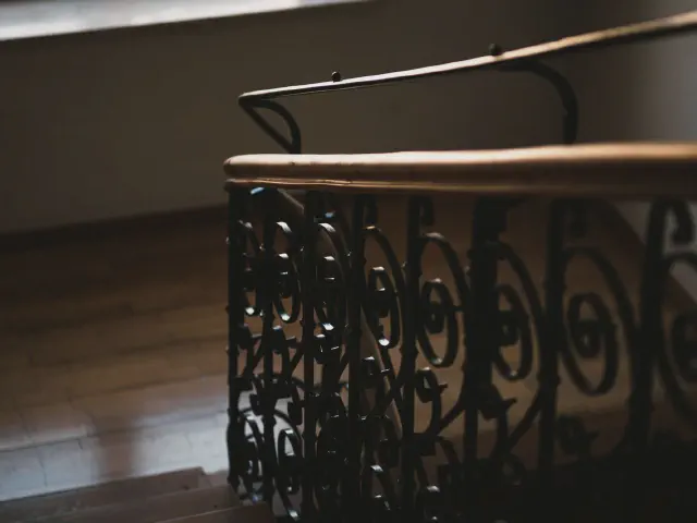 Zakręcona balustrada z czarnego metalu w gęste, kręcone ornamenty zwieńczona drewnianą poręczą, w półmroku