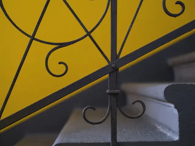 Zbliżenie balustrady z czarnego metalu z kręconymi ornamentami na tle żółtej ściany i stopni z szarą wykładziną