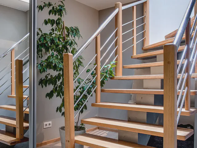 Ażurowe drewniane schody z balustradą z cienkich, poziomych prętów stalowych mocowanych do drewnianych słupków, obok roślina