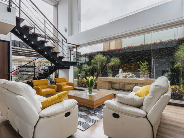 Wysoki salon z przeszkloną ścianą, w tle czarne stalowe schody z balustradą z poziomych prętów prowadzące na antresolę