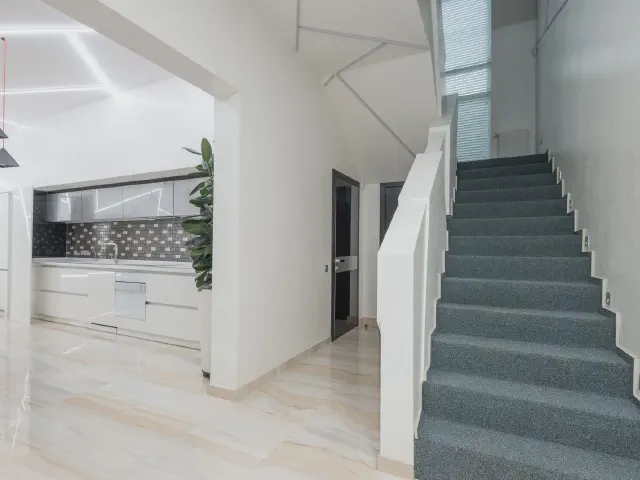 Korytarz przy jasnej kuchni, szare schody z wykładziną ograniczone białą, pełną ścianką zamiast balustrady