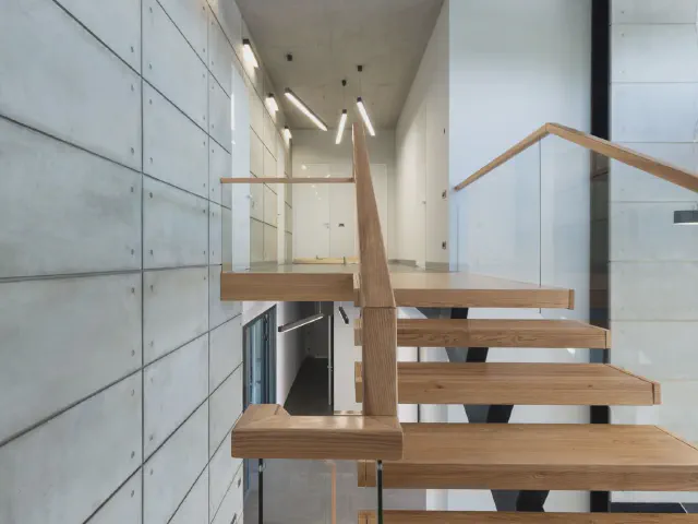 Drewniane stopnie wspornikowe przy ścianie z płyt betonowych, zabezpieczone bezramową szklaną balustradą z drewnianą poręczą