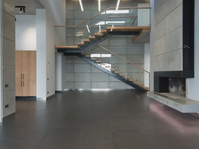Przestronne wnętrze z ciemną posadzką i kominkiem, przy ścianie schody wspornikowe ze szklaną balustradą