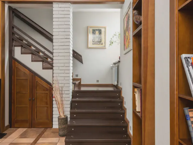 Drewniana szafka z drzwiczkami w przestrzeni pod schodami, obok ceglany fragment ściany i regał z książkami, ciemne drewniane stopnie