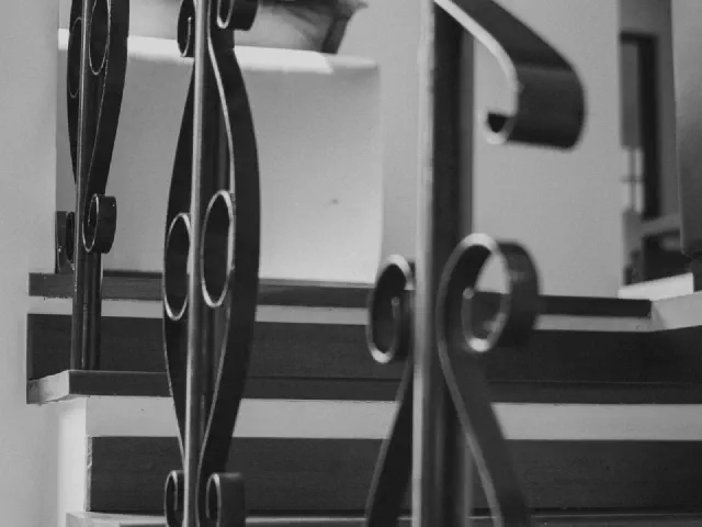 Zbliżenie czarnych, kutych tralek z ornamentem przy stopniach z czarnymi nakładkami na tle białej ściany