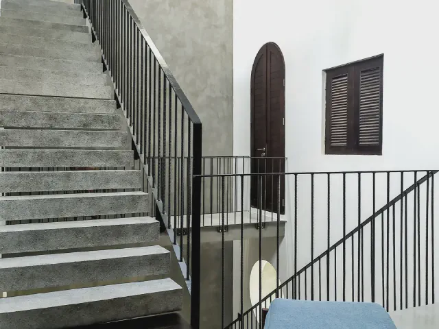 Szare, surowe schody betonowe z czarną metalową balustradą z pionowych prętów, obok biała ściana z łukowymi drzwiami