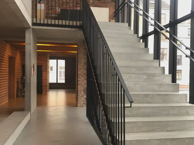 Szare betonowe schody z czarną metalową balustradą w industrialnym wnętrzu z ceglaną ścianą i przeszkleniem