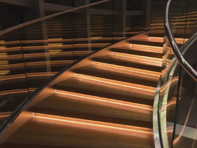 Kręte schody ze szklaną balustradą, w stopniach wbudowane pasy światła LED w ciepłej barwie