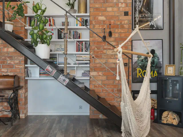 Loft z odsłoniętą ceglaną ścianą, czarnymi stalowymi schodami policzkowymi, regałem z książkami pod biegiem i hamakiem