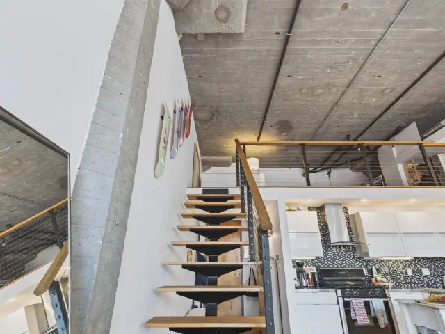 Loft z betonowym stropem i słupem, schody z drewnianymi stopniami na centralnej belce prowadzą na antresolę nad kuchnią