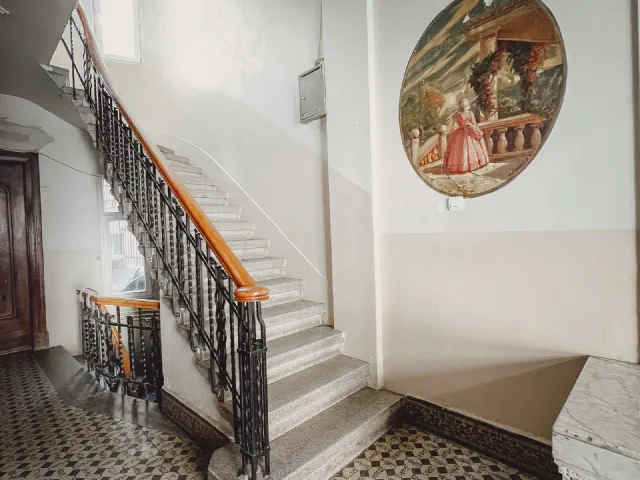 Białe kamienne schody z czarną kutą balustradą i drewnianym pochwytem, na posadzce wzorzyste płytki cementowe, na ścianie malowany talerz