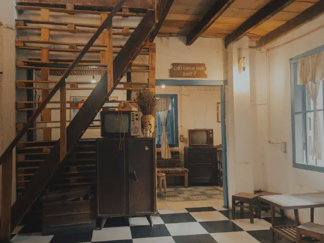 Wnętrze w stylu retro z czarno-białą szachownicą na podłodze, prostymi drewnianymi schodami przy ścianie z desek, belkowym stropem i starym telewizorem na szafce