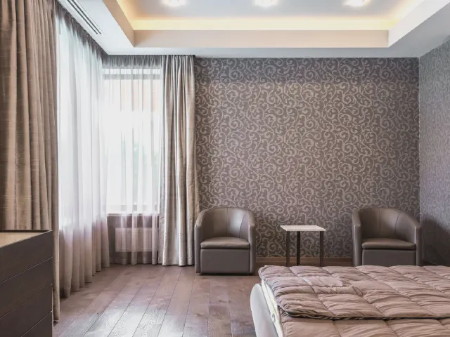 Sypialnia z tapetą w ornament na ścianie, dwoma skórzanymi fotelami przy małym stoliku, długimi zasłonami i drewnianą podłogą