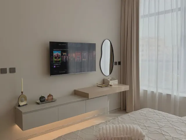 Telewizor na beżowej ścianie nad długą wiszącą szafką z podświetleniem, obok owalne lustro i blat toaletki, zasłony przy dużym oknie