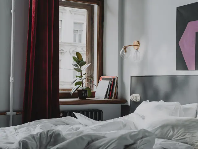 Sypialnia z szarymi ścianami i szarym zagłówkiem, bordowymi aksamitnymi zasłonami przy oknie, białą pościelą i mosiężnym kinkietem