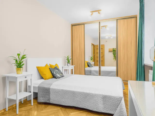 Sypialnia z białym łóżkiem, szarą narzutą i żółtymi poduszkami, szafą z lustrzanymi i drewnianymi drzwiami, parkietem w jodełkę i białą szafką nocną z rośliną