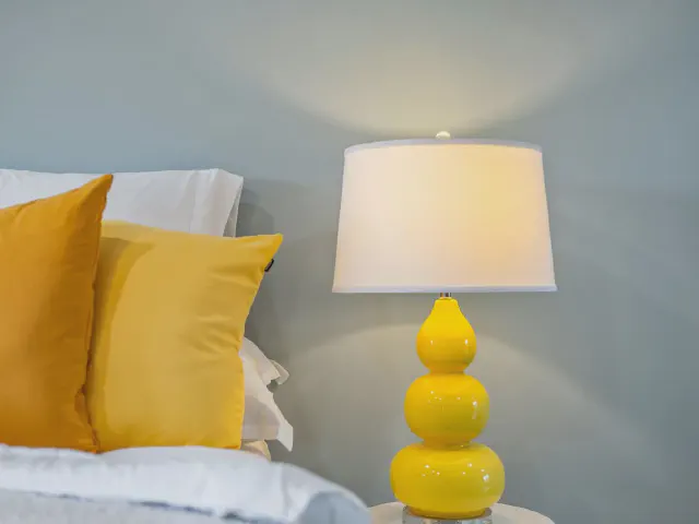 Łóżko z białą pościelą i dwiema musztardowymi poduszkami, obok lampka nocna z żółtą ceramiczną podstawą na tle szaroniebieskiej ściany