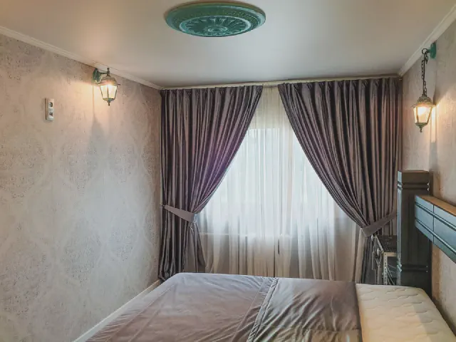 Sypialnia z tapetą w ornament, kinkietami w stylu latarni i śliwkowymi zasłonami upiętymi ozdobnymi opaskami po bokach okna z białą firaną