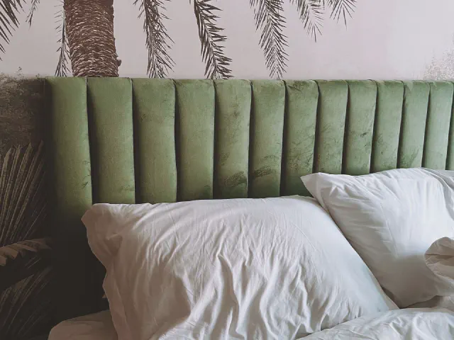 Zbliżenie łóżka z zielonym welurowym wezgłowiem w pionowe przeszycia na tle fototapety z liśćmi palm na jasnoróżowym tle