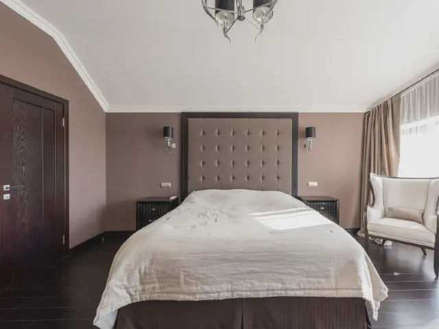 Wysoki panel tapicerowany w szarobeżowej tkaninie z pikowaniem guzikami, sięgający sufitu, za łóżkiem z jasną narzutą, kinkiety po bokach i fotel przy oknie