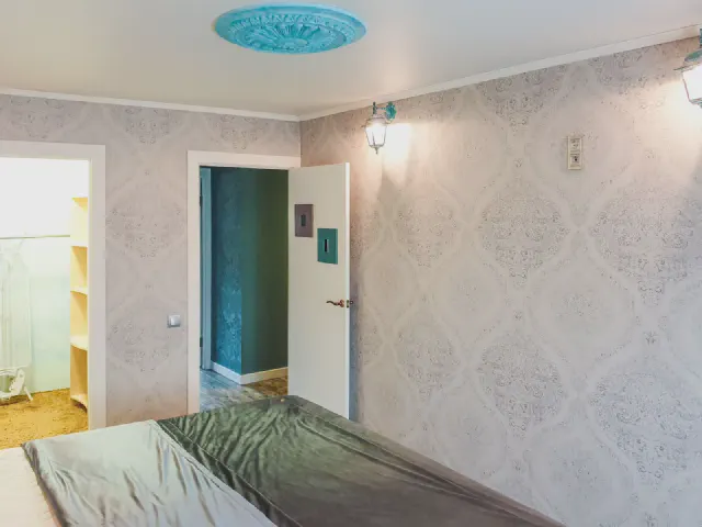 Jasna tapeta w delikatny, jasnoszary ornament na wszystkich ścianach sypialni, łóżko z zieloną narzutą i otwarte drzwi do garderoby