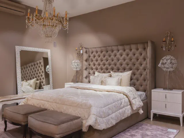 Sypialnia glamour z wysokim, pikowanym zagłówkiem w kolorze taupe, kryształowym żyrandolem, lustrem w białej ramie i dwiema ławami w nogach łóżka