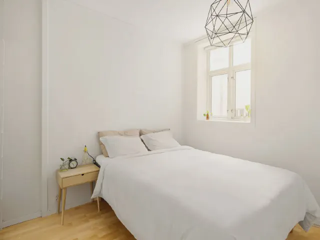 Białe łóżko z jasną drewnianą szafką nocną i geometryczną lampą wiszącą przy oknie