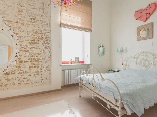 Sypialnia z białym metalowym łóżkiem z ornamentem, ścianą z bielonej cegły, rzymską roletą i kolorowym żyrandolem