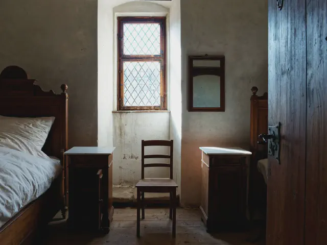 Sypialnia w starym domu z kamiennymi ścianami, ciemnym drewnianym łóżkiem, małym oknem z witrażowymi szybkami i drewnianym krzesłem