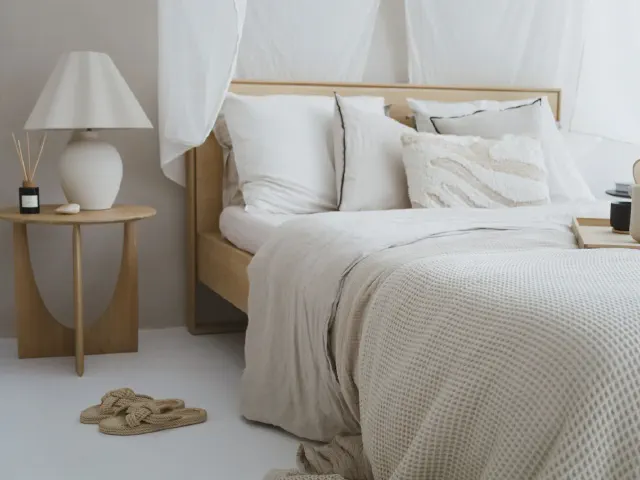 Drewniane łóżko z lnianą pościelą i narzutą z wafelkowej bawełny, biała ceramiczna lampa na okrągłym drewnianym stoliku